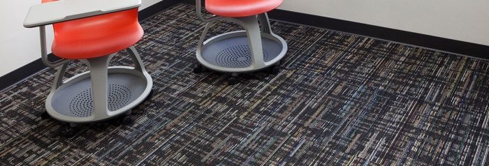 Studio shot of a two classroom desk chairs, resting atop Relativity carpeting. Each desk chair uses red plastic on the chair and grey desk top. They have a full platform for the feet, with wheels.