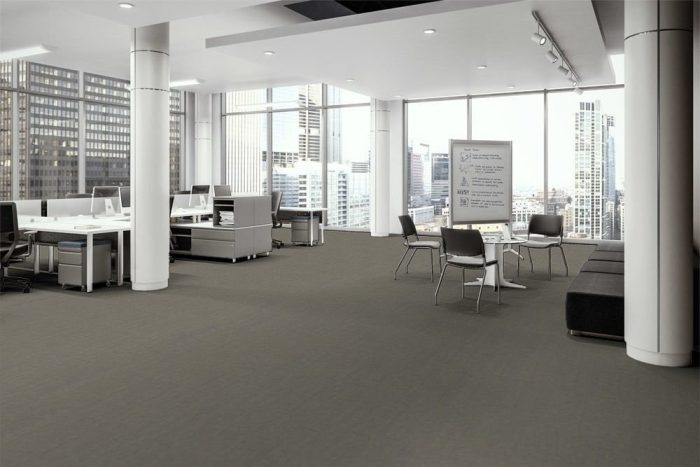 Transit flooring in the open office. At one side are occupied work desks with attached credenzas. A portable white board is placed to the side of one round cafe table, with instructions written upon it. The full height windows look out to the city.